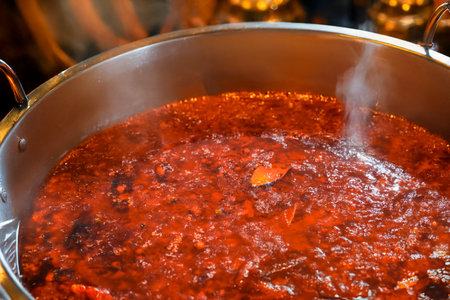 A stainless steel pot filled with rich red chili broth and floating herbs served on a wooden table.の写真素材