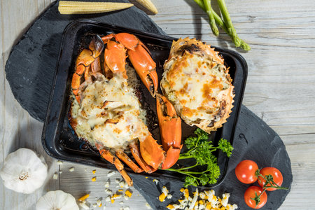 A black tray with two crabs baked in creamy cheese topping surrounded by fresh vegetables and garlic cloves.の写真素材