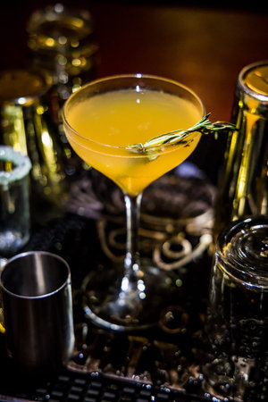 A yellow-orange cocktail in a coupe glass garnished with rosemary. Bar tools and lighting create a...