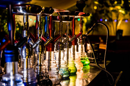 A row of hookahs with glass bases and metal stems arranged on a counter. Warm lighting and...