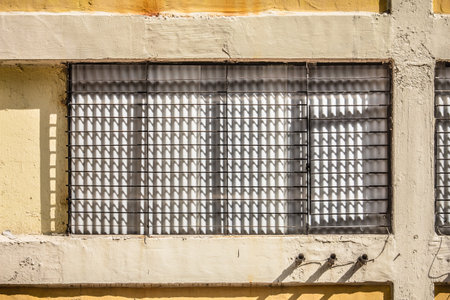 A building facade with a window grille casting shadows, pipes, and aged yellow wall, showing...