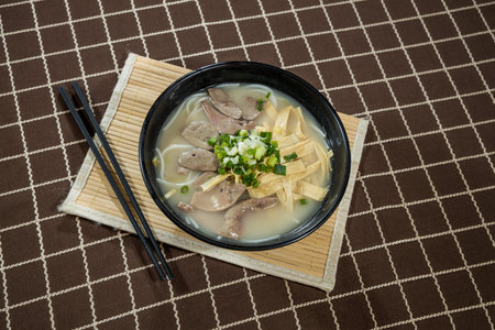 A black bowl of clear broth with slices of pork liver, tofu strips, and chopped green onions, served...