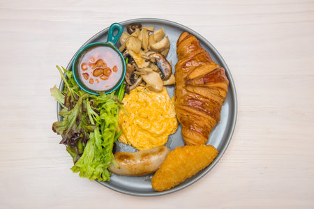 A round plate featuring croissant, scrambled eggs, sausage, salad, and mushrooms.