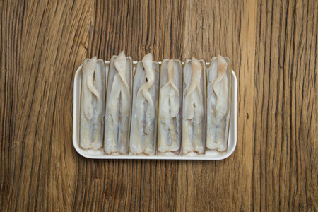 A small white tray displays several fresh razor clams lined up uniformly on a wood grain board. Their translucent flesh and smooth shells convey freshness, suitable for steaming, quick stir fry, or grilling. Minimal styling emphasizes the ingredient itself.の写真素材
