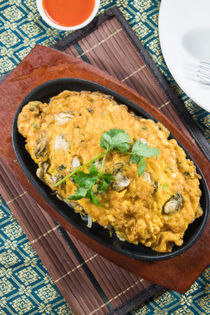Close view of an oyster omelette served on a hot iron plate with a side of bright red sauce. Puffy eggs, seared edges, and fresh herbs convey heat and aroma. A patterned cloth adds cultural context. Great for street food features and night market themes.の写真素材