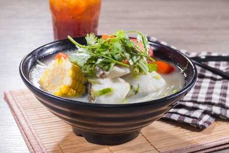 A black ceramic bowl filled with clear broth, fish fillets, corn, carrots, and fresh herbs served on...