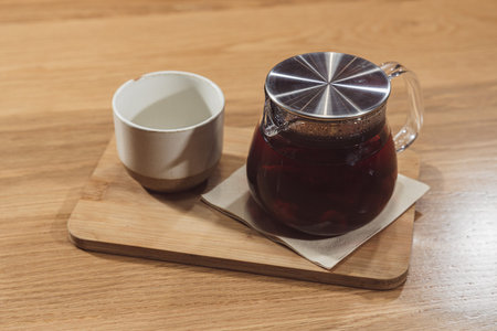 A clear teapot filled with dark hot tea sits on a wooden board beside a ceramic cup and napkin, a cozy beverage moment on a warm wood table.
