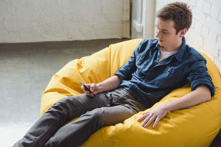 Attentive young man sitting in yellow bean bag chair and using black smartphone indoorsの写真素材