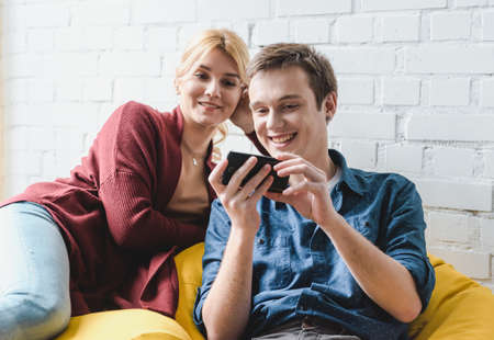Smiling young couple sitting on yellow bean bag chair and looking at black smartphone indoorsの写真素材