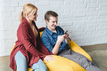 Smiling young couple sitting on yellow bean bag chair and looking at black smartphone indoorsの写真素材