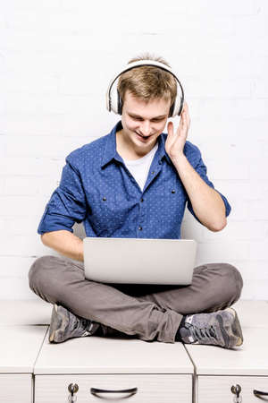 Handsome young man in white headphones sitting on chest of drawers and using laptop against white brick wall backgroundの写真素材