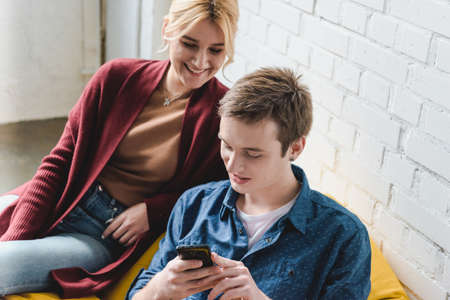 Smiling young couple sitting on yellow bean bag chair and looking at black smartphone indoorsの写真素材