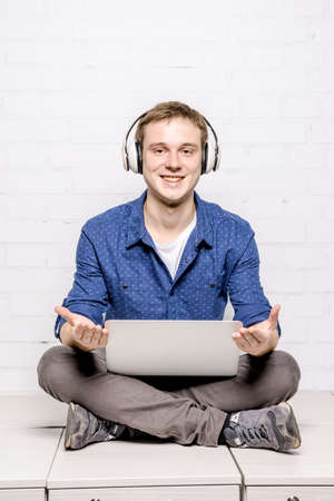 Handsome young man in white headphones sitting on chest of drawers and using laptop against white brick wall backgroundの写真素材