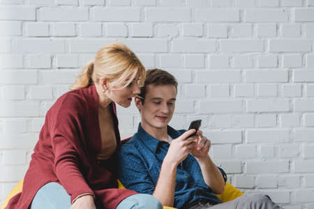 Smiling young couple sitting on yellow bean bag chair and looking at black smartphone indoorsの写真素材