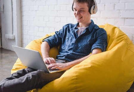 Smiling young man in white headphones sitting in yellow bean bag chair and using laptop indoorsの写真素材