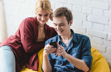 Smiling young couple sitting on yellow bean bag chair and looking at black smartphone indoorsの写真素材