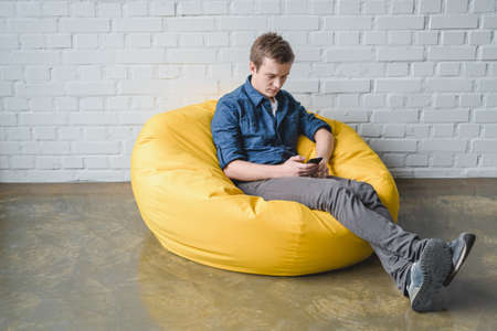 Attentive young man sitting in yellow bean bag chair and using black smartphone indoorsの写真素材