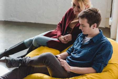 Smiling young couple sitting on yellow bean bag chair and looking at black smartphone indoorsの写真素材