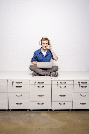 Handsome young man in white headphones sitting on chest of drawers and using laptop against white...