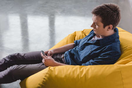 Attentive young man sitting in yellow bean bag chair and using black smartphone indoorsの写真素材