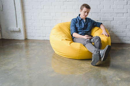 Attentive young man sitting in yellow bean bag chair and using black smartphone indoorsの写真素材