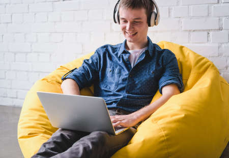 Smiling young man in white headphones sitting in yellow bean bag chair and using laptop indoorsの写真素材