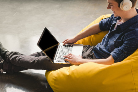 Smiling young man in white headphones sitting in yellow bean bag chair and using laptop indoors