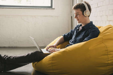 Smiling young man in white headphones sitting in yellow bean bag chair and using laptop indoorsの写真素材