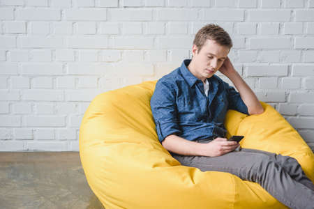 Attentive young man sitting in yellow bean bag chair and using black smartphone indoorsの写真素材