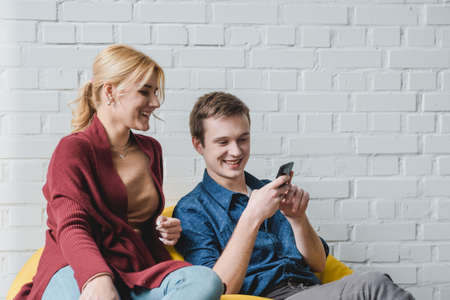 Smiling young couple sitting on yellow bean bag chair and looking at black smartphone indoorsの写真素材