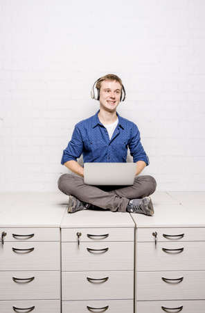 Handsome young man in white headphones sitting on chest of drawers and using laptop against white brick wall backgroundの写真素材