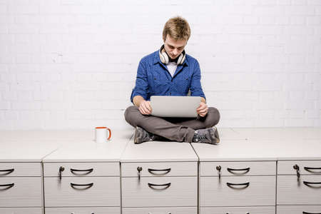 Handsome young man in white headphones sitting with coffee cup on chest of drawers and holding laptop against white brick wall backgroundの写真素材