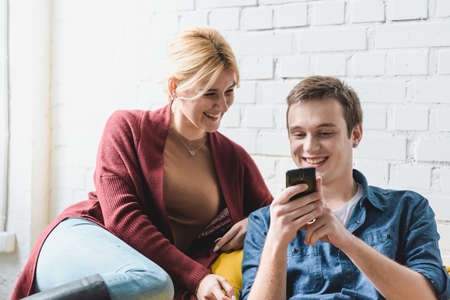 Smiling young couple sitting on yellow bean bag chair and looking at black smartphone indoorsの写真素材
