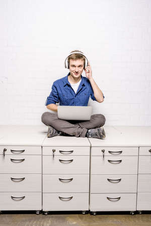 Handsome young man in white headphones sitting on chest of drawers and using laptop against white brick wall backgroundの写真素材