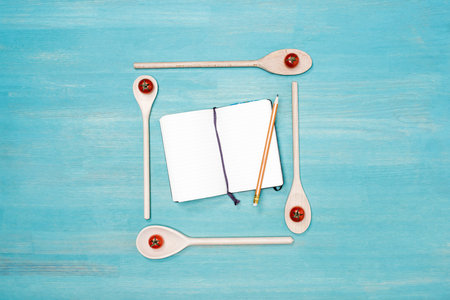 Top view of wooden spoons with cherry tomatoes and blank open cookbook with pencil on table