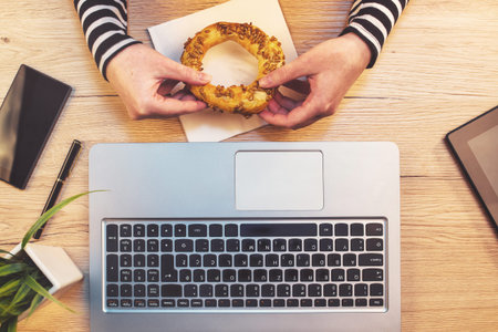 Woman eating sesame bagel for breakfast in office, top view of female hands holding baked snack on...