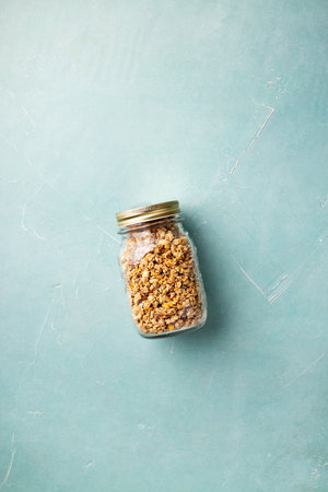 Home granola in a glass jar on blue concrete background.Healthy vegan snack. Flat layの写真素材