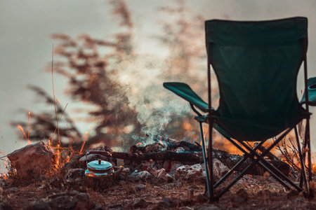 Conceptual Photo of an Autumnal Peaceful Recreation. Empty Camp Chair near Teapot in the Campfire....