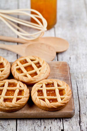 Fresh baked tarts with marmalade or apricot jam filling and on cutting board and kitchen bakery utensil on on rustic wooden table background.の写真素材