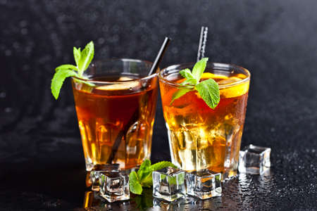 Two glasses with cold traditional iced tea with lemon, mint leaves and ice cubes on wet black background.の写真素材