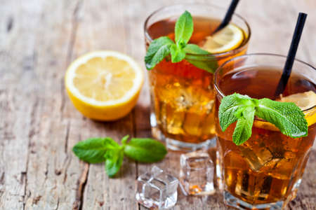 Cold iced tea with lemon, mint leaves and ice cubes in two glasses on rustic wooden table background. With copy space.の写真素材