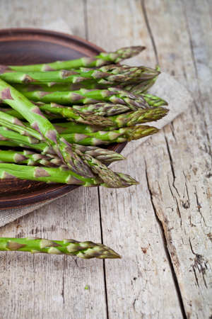 Fresh raw garden asparagus closeup on brown ceramic plate and linen napkin on rustic wooden table background. Green spring vegetables. Edible sprouts of asparagus. With copy space.の写真素材