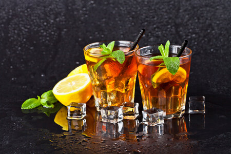 Two glasses with cold traditional iced tea with lemon, mint leaves and ice cubes on wet black background.の写真素材