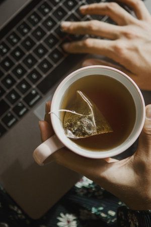 Woman holding a cup of tea while using a laptopの素材