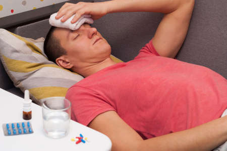 Man with a headache lying on the sofa. holds a compress on his head. pills and a glass of water on the table.の写真素材