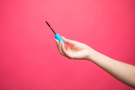 Close up of womans hand holding an eyelash brush mascara on pink background