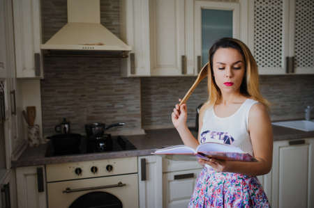 young woman is cooking in the vintage kitchen with joy. She is standing and holding a book of recipe. The lady is touching a wood spoon to her face and dreaming. She is smilingの写真素材
