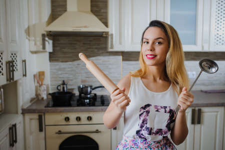 young woman is cooking in the kitchen with joy. She is standing and holding rolling pin and spoonの写真素材