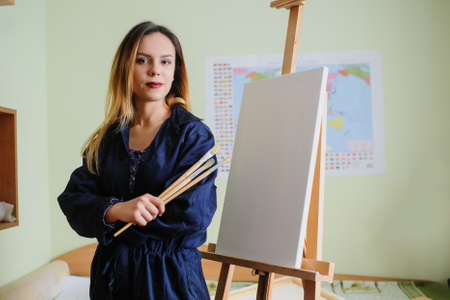 Attractive young woman artist begins to draw a new paint on a  wooden easel, holds brush.の写真素材