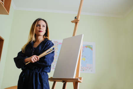 Attractive young woman artist begins to draw a new paint on a  wooden easel, holds brush.の写真素材